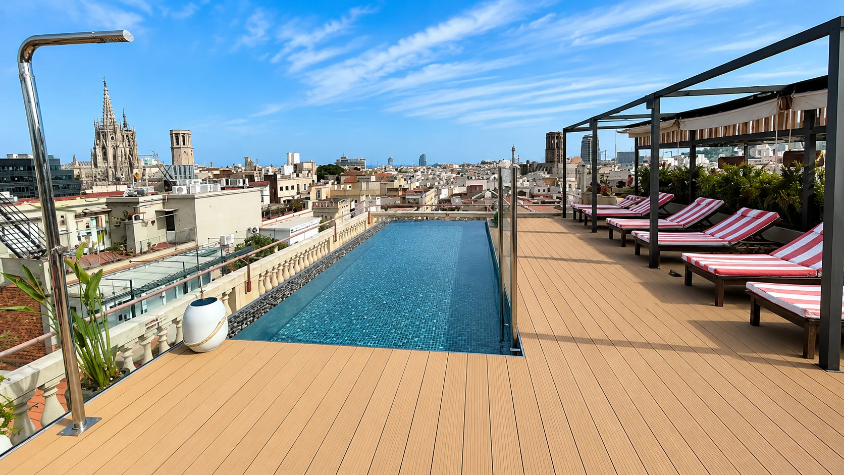 Rooftop infinity pool and striped sun loungers at the Kimpton hotel in Barcelona's Gothic Quarter, overlooking the city rooftops and the spire of Barcelona Cathedral under a blue sky